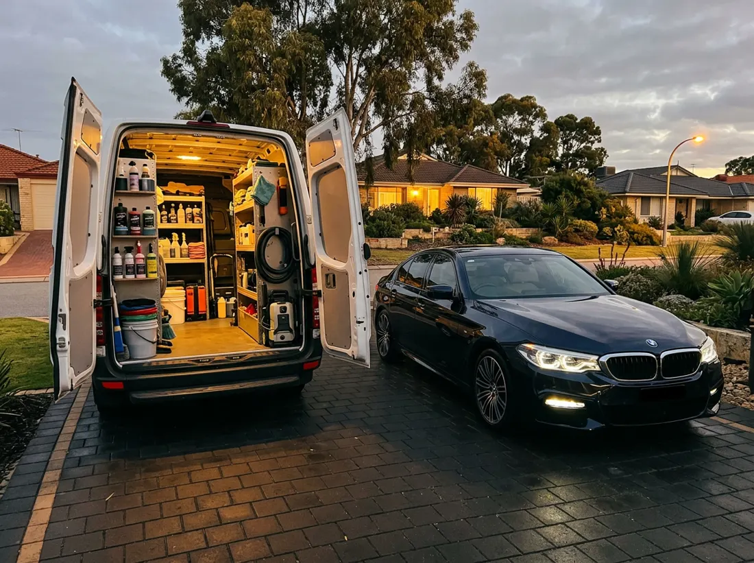 Mobile detailing van with its rear doors open beside a gleaming dark sedan with clear headlights on a Perth driveway at dusk